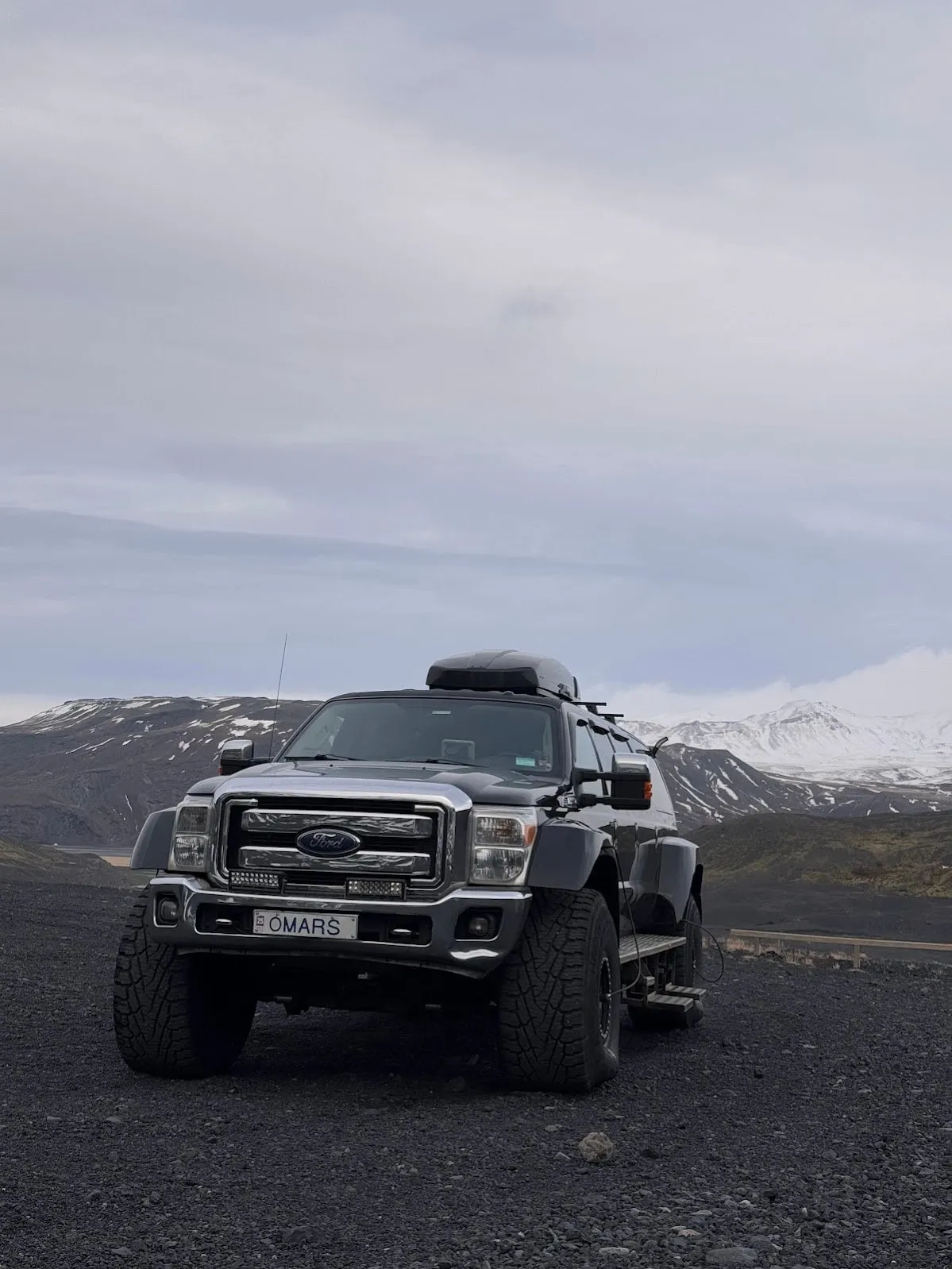 Lifted black Ford super-jeep with massive off-road tyres parked on black volcanic gravel, snow-capped mountains in the distance