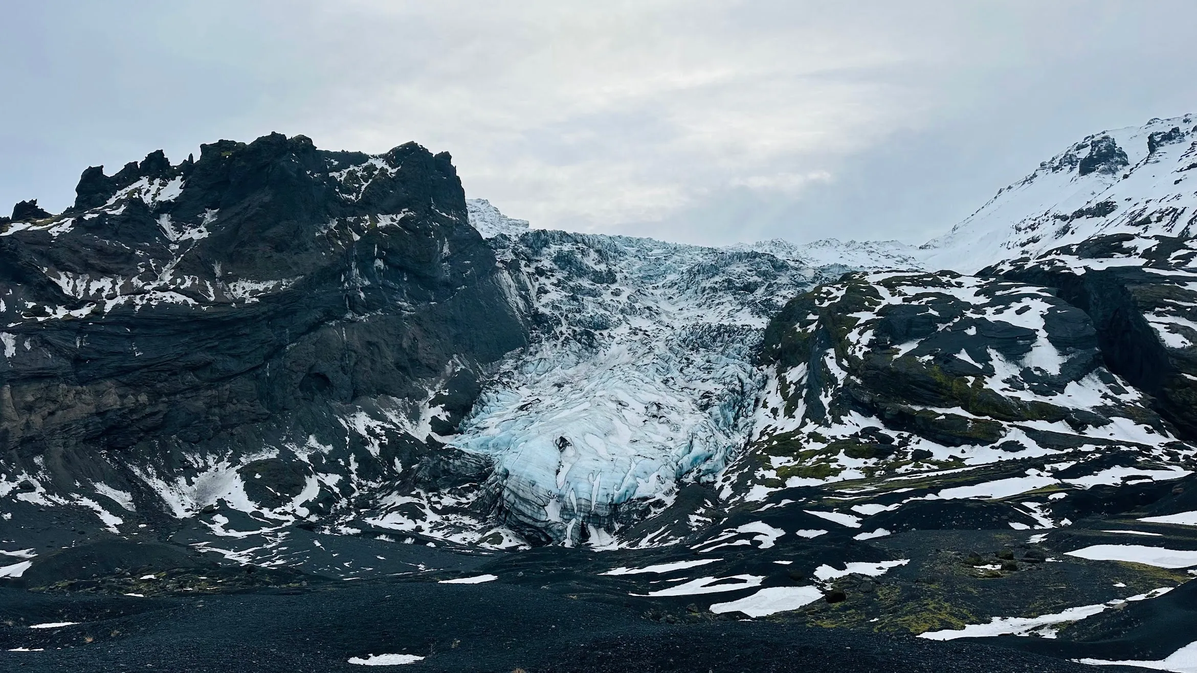 Pale blue glacier tongue spilling between dark volcanic rock walls down to a foreground of black gravel