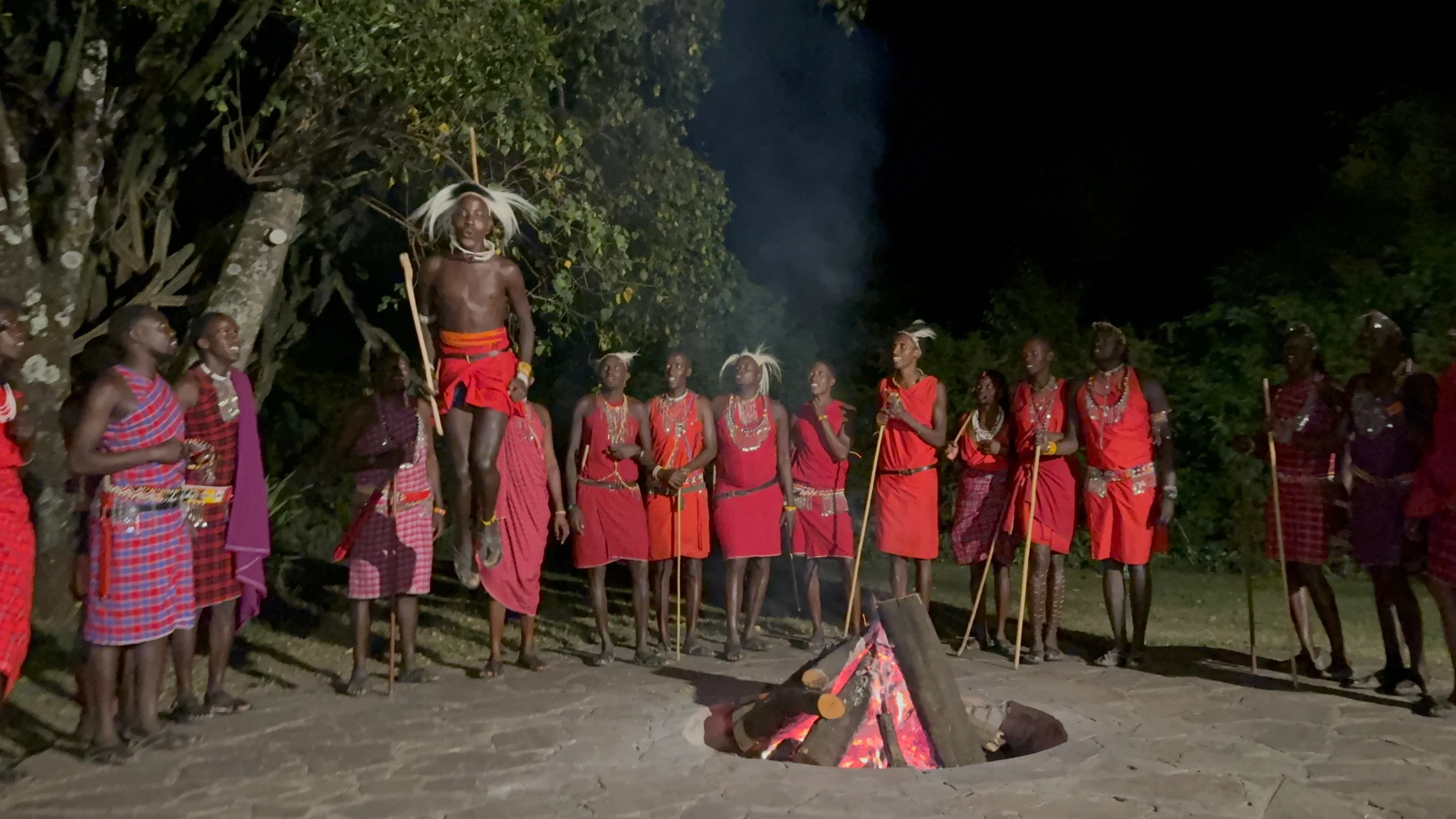 Maasai warriors in red shukas around a fire at night, one warrior mid-jump in traditional adumu dance