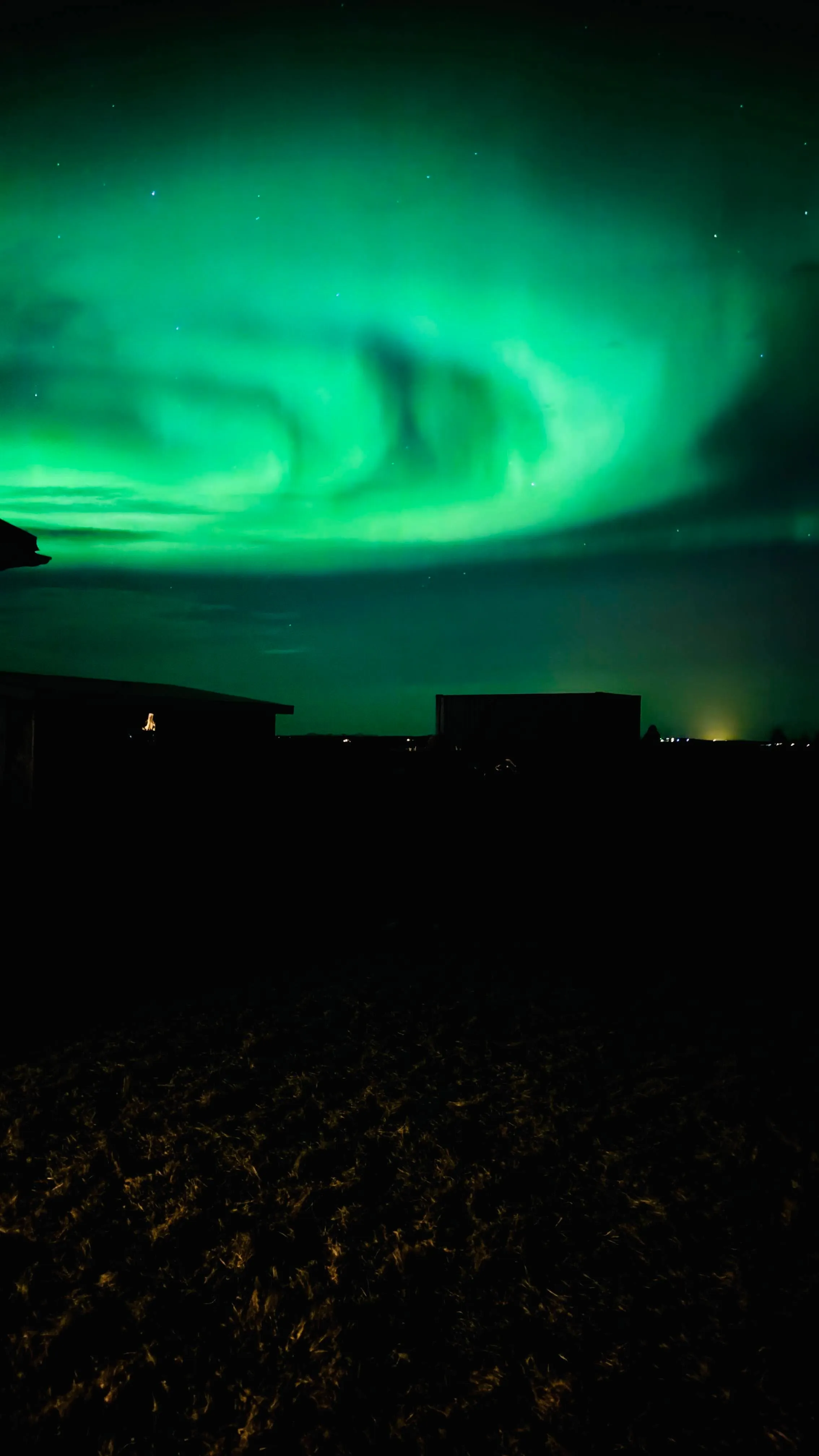 Wide green aurora arc over silhouetted farm buildings near Hella, with a small bonfire glowing on the left and a cabin window lit in the distance