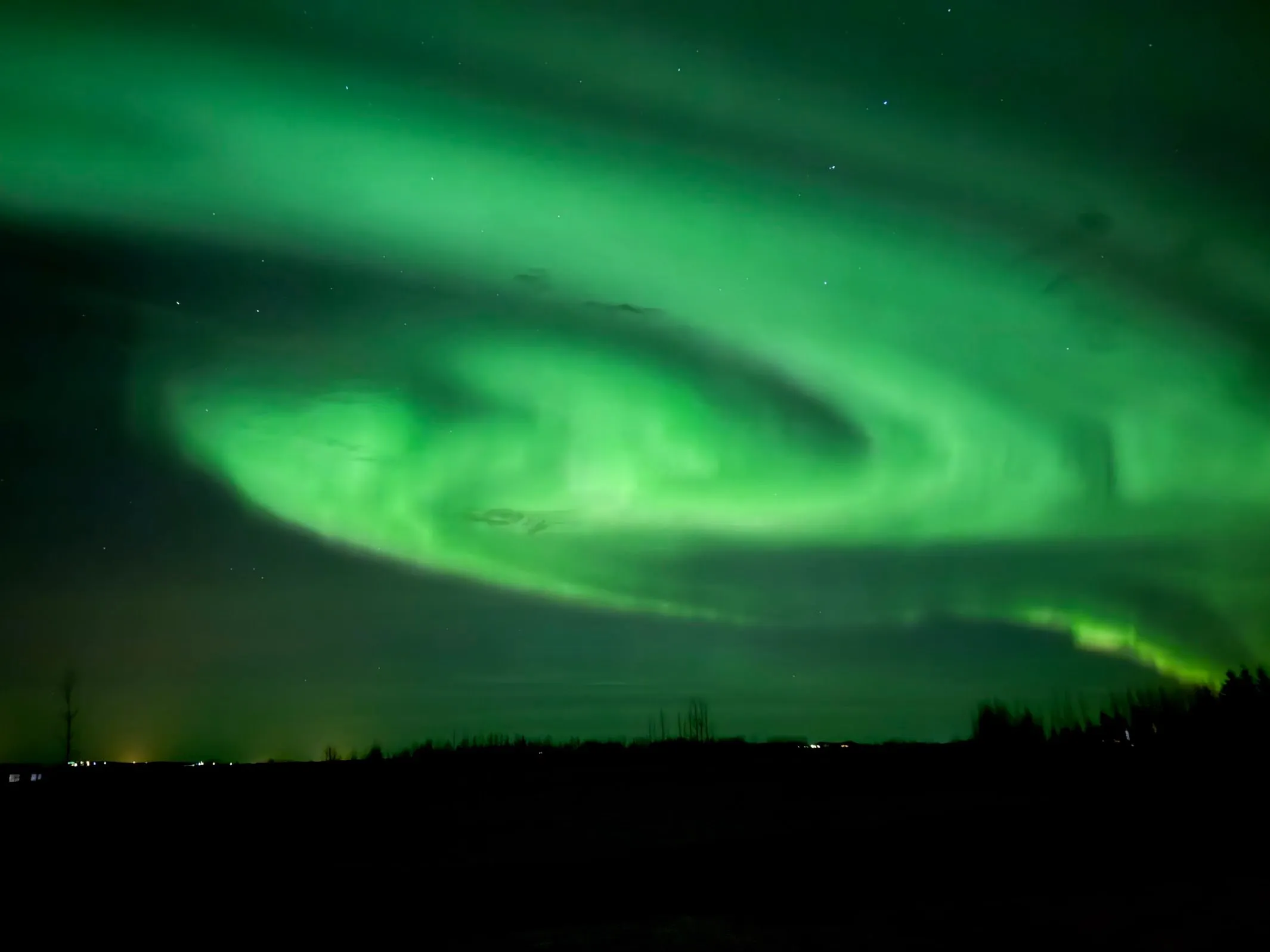 Brilliant green aurora swirling in a tight spiral over a silhouetted treeline, scattered stars visible in the dark sky over Hella