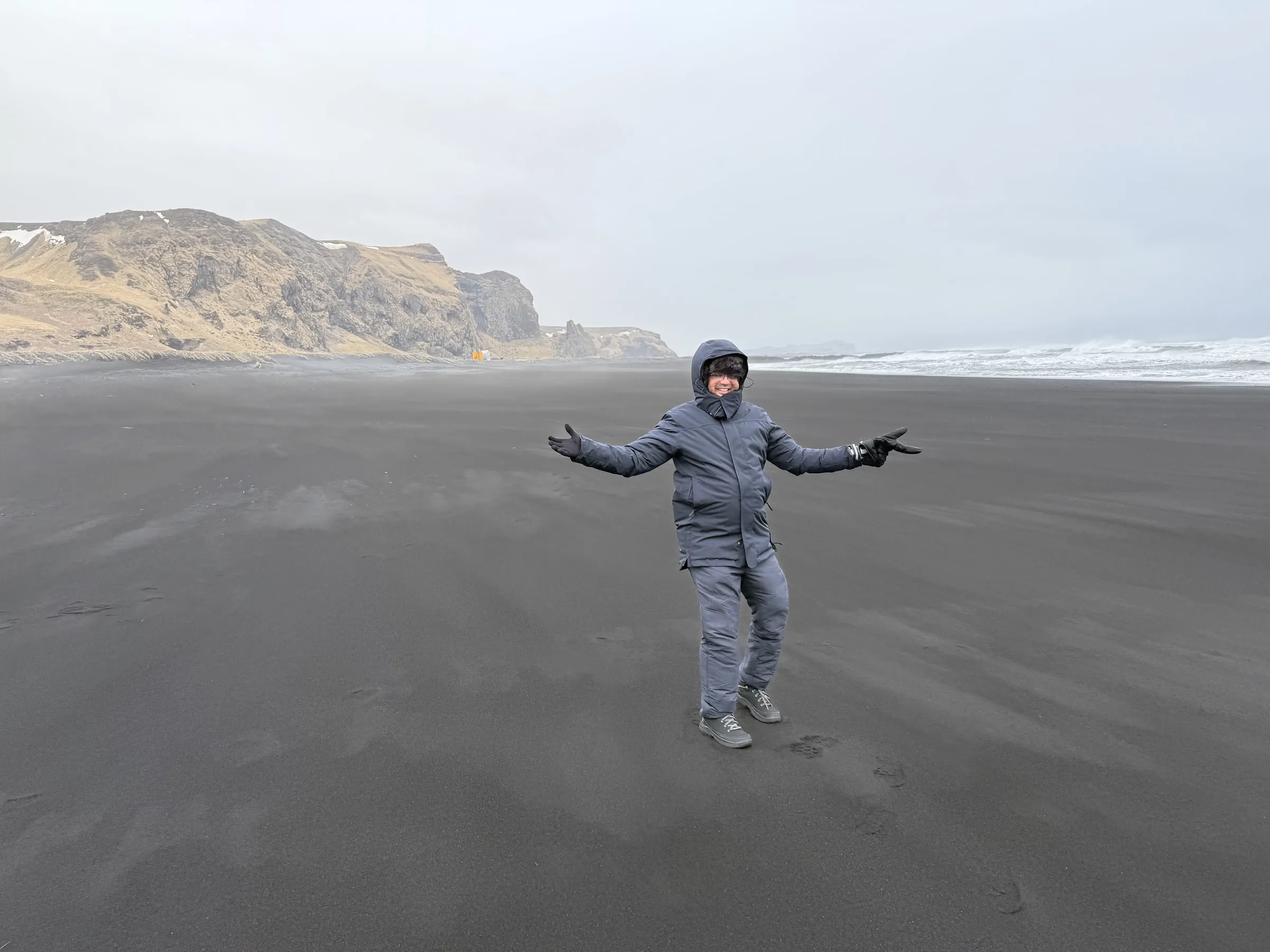 Person in a grey insulated suit standing arms wide on the black volcanic sand of Reynisfjara, foamy Atlantic surf to the right, cliffs to the left