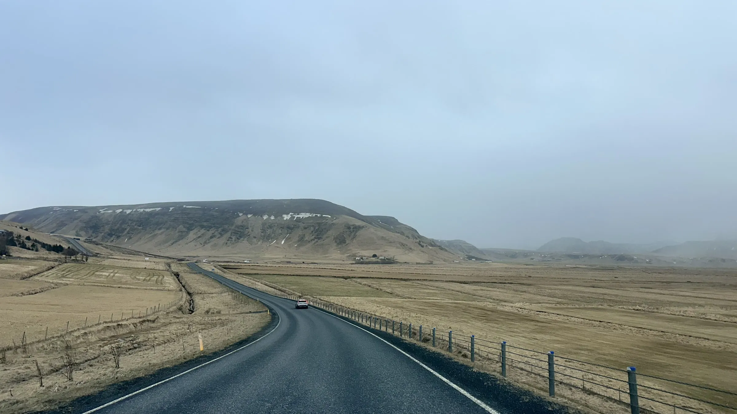 Dark asphalt of Route One curving through pale tussock plains toward a flat-topped ridge under a hazy Icelandic sky, a single car in the distance