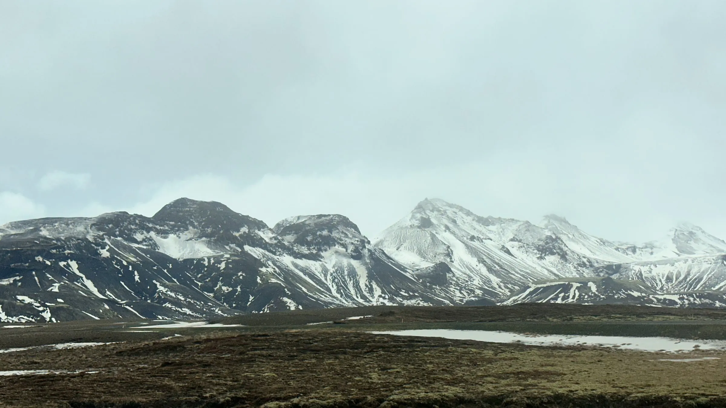 Snow-streaked dark mountains rising over a marshy plain with patches of ice and grass, under heavy grey clouds