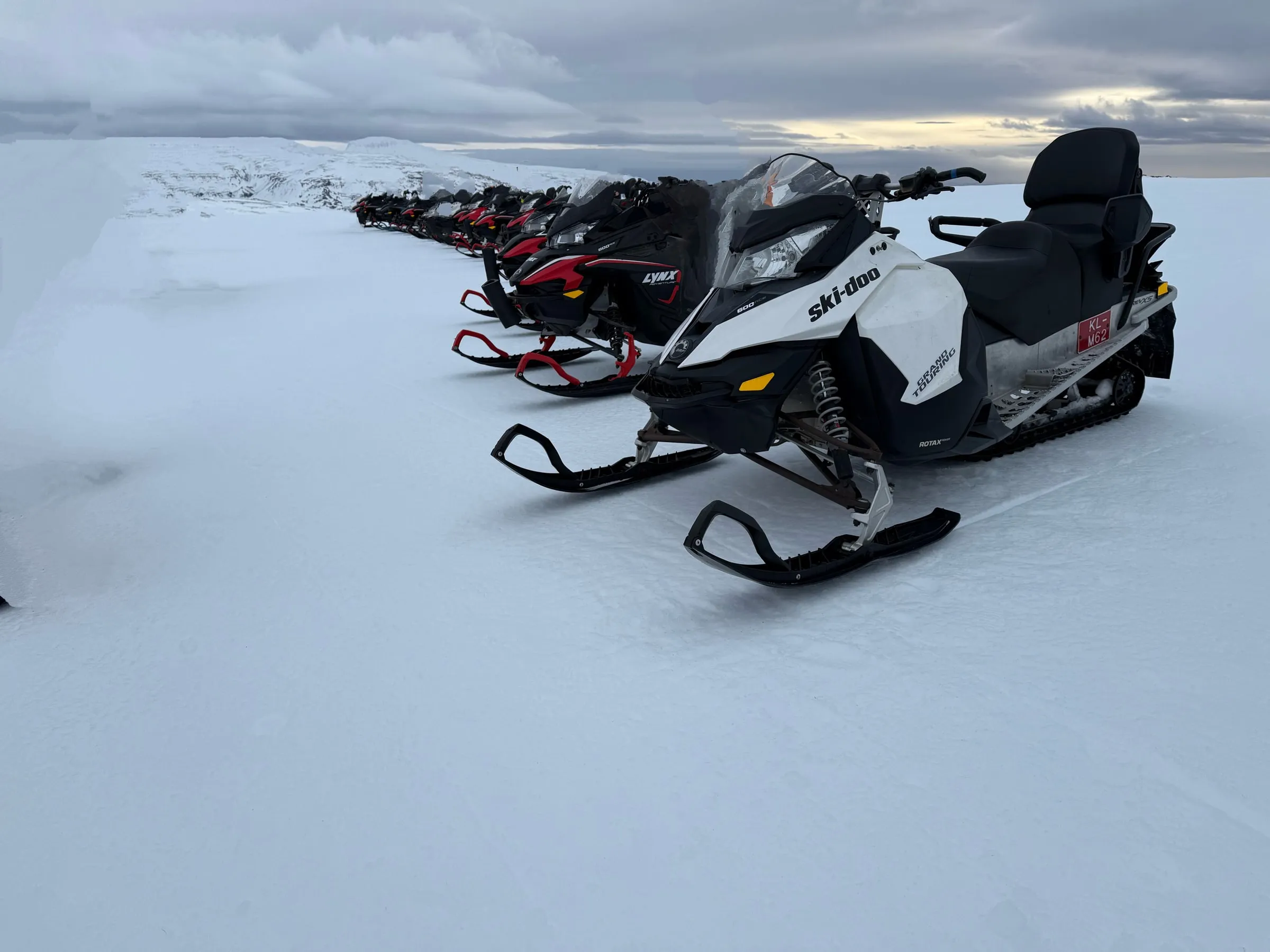 Row of Ski-Doo snowmobiles parked single-file on a glacier, sun breaking through clouds on the horizon