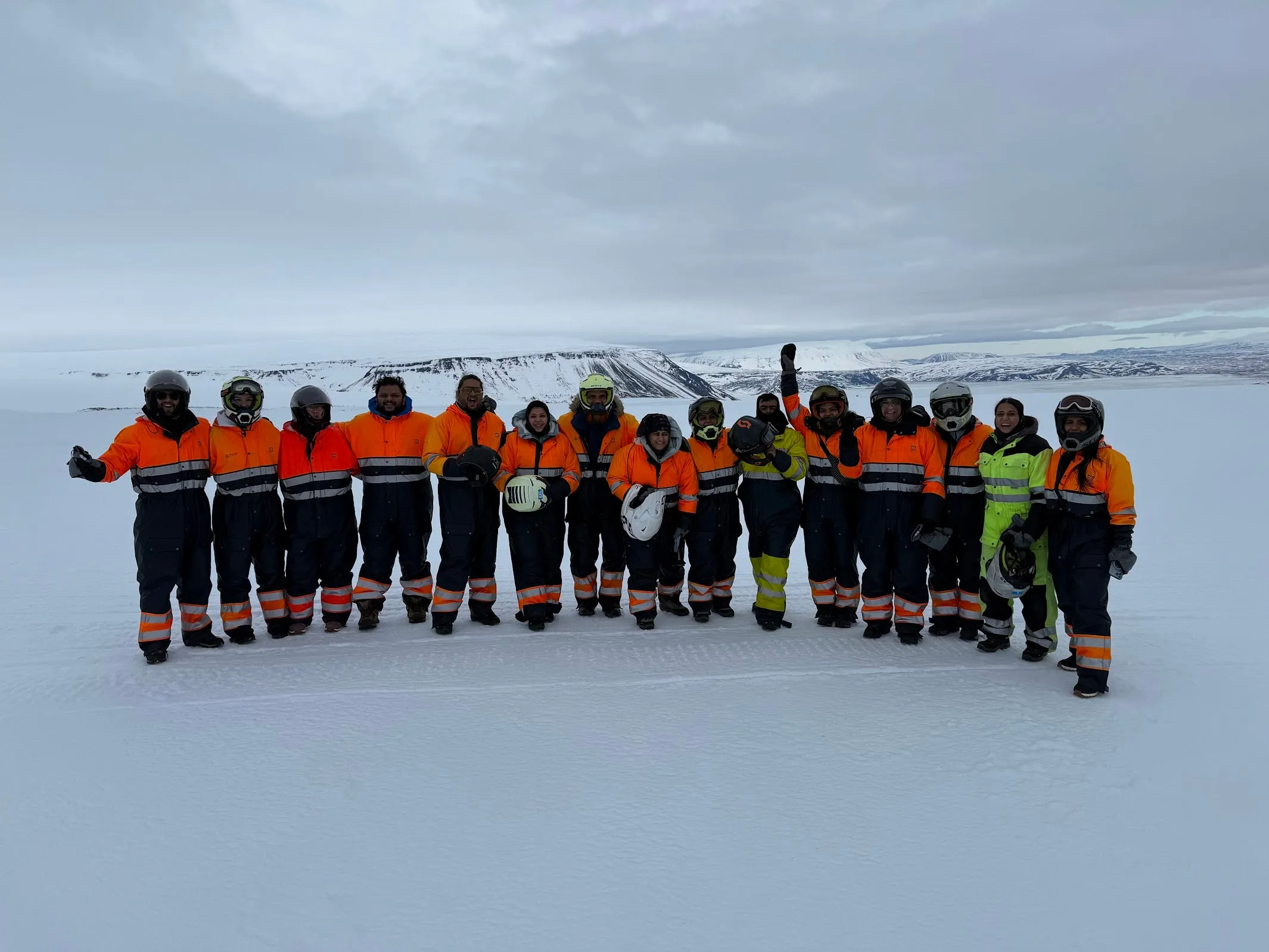 Fifteen people in orange and black snow gear lined up arm-in-arm across a glacier, helmets on, snow-capped ridge behind them