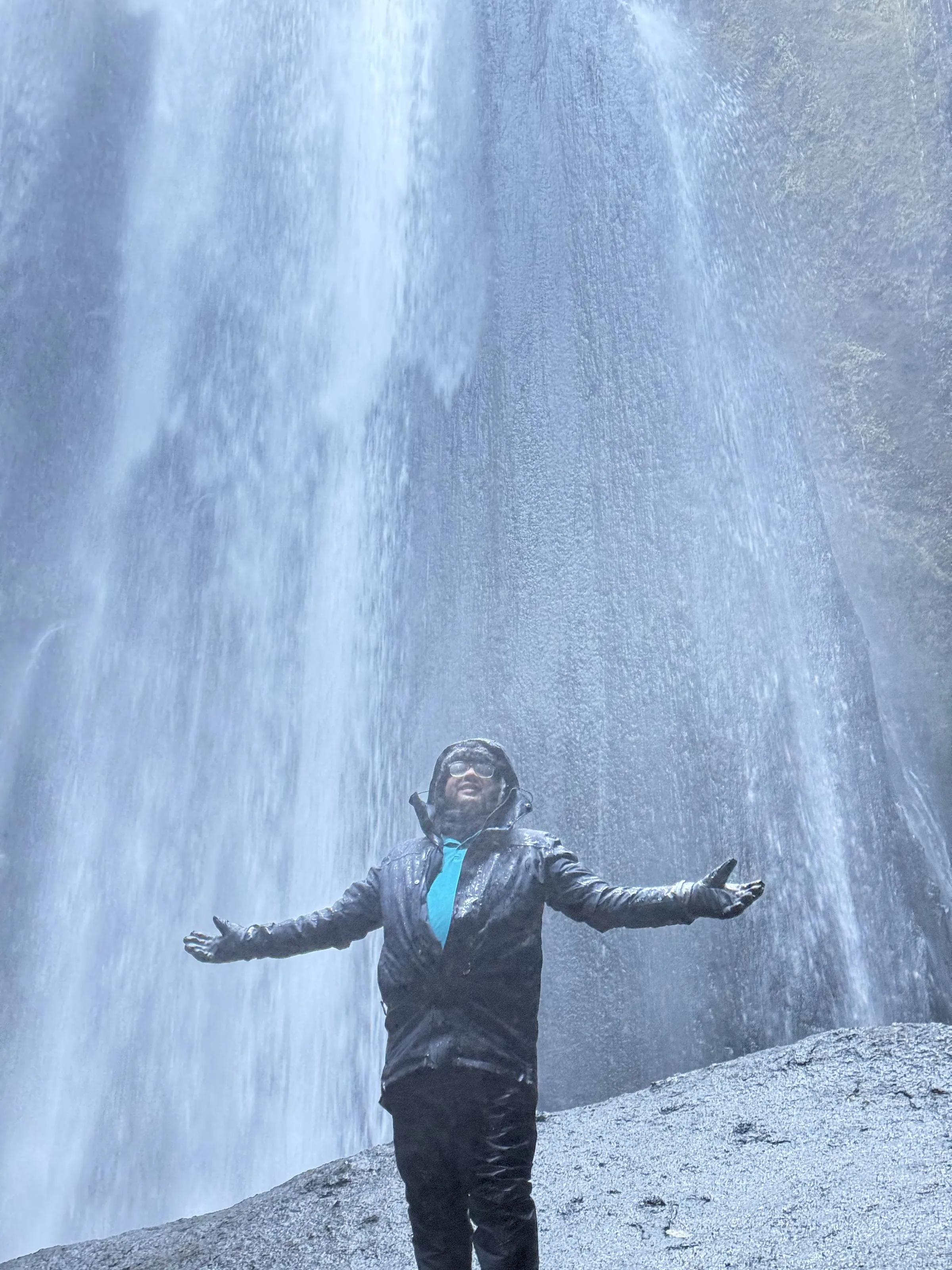 Person in a soaked black jacket standing arms wide directly under a thundering Icelandic waterfall, spray filling the frame