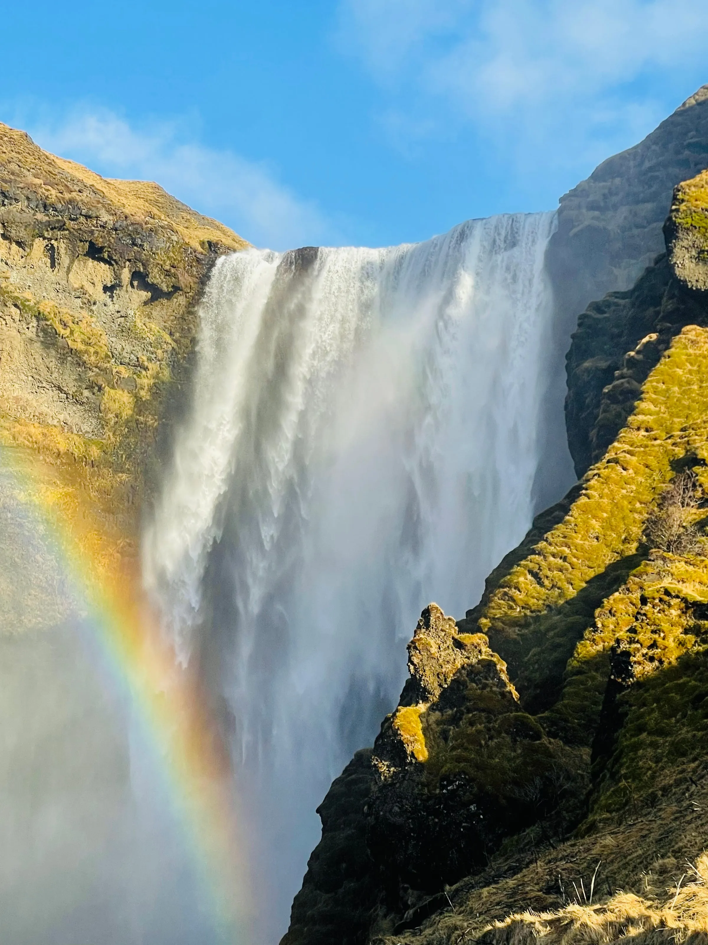 Tall waterfall plunging down a mossy cliff with a vivid rainbow forming in the spray on the left side of the frame